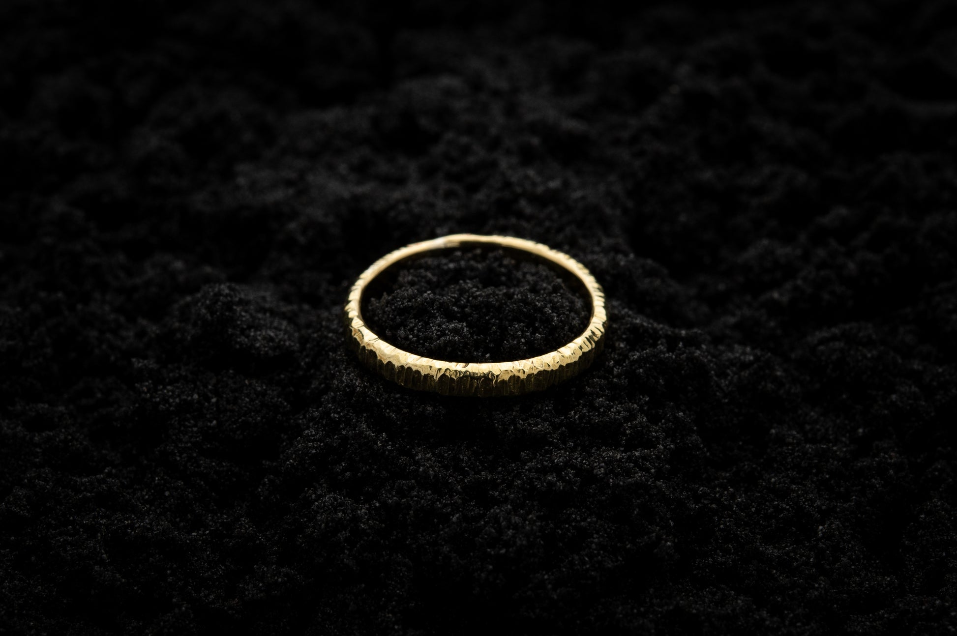 A slim, yellow gold wedding ring sits atop black sand to contrast its warm, luscious glow. There are hammered striations around the band like the suns rays.