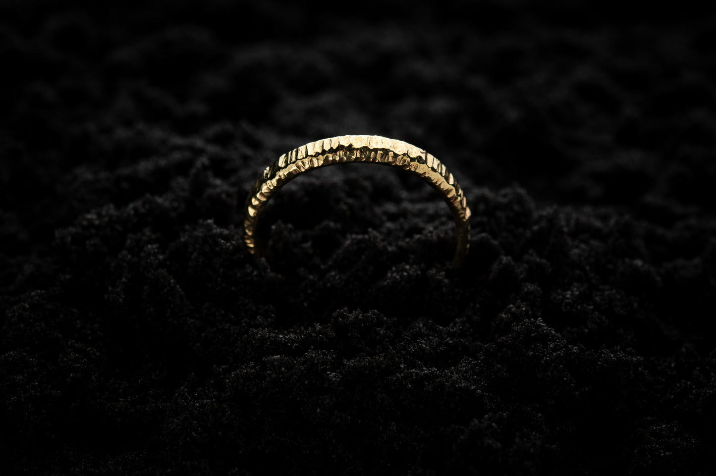 A slim, yellow gold wedding ring sits in black sand to contrast its warm, luscious glow. There are hammered striations around the band like the suns rays. It is half submerged in the sand and breaks through the surface like a golden rainbow.