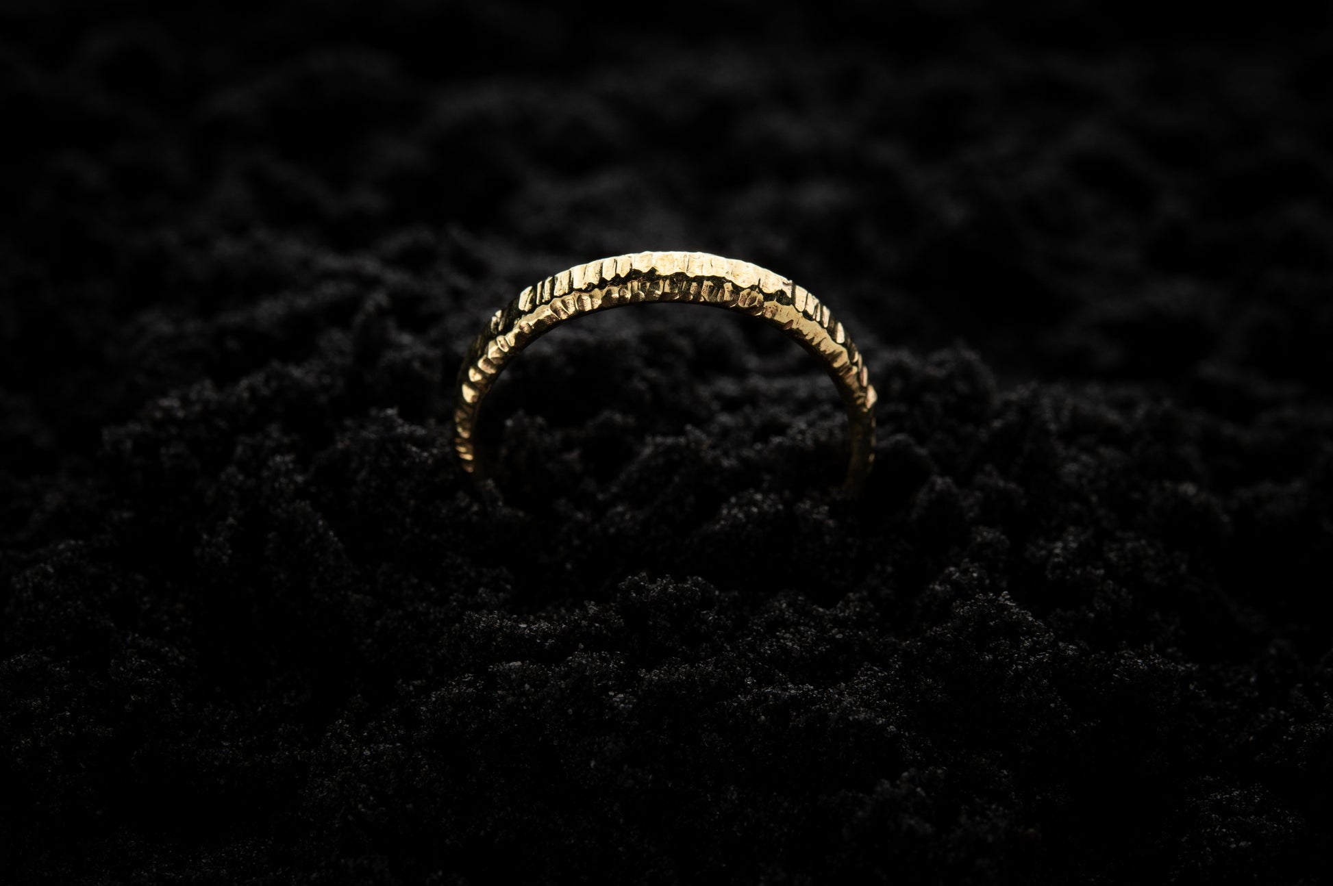 A slim, yellow gold wedding ring sits in black sand to contrast its warm, luscious glow. There are hammered striations around the band like the suns rays. It is half submerged in the sand and breaks through the surface like a golden rainbow.