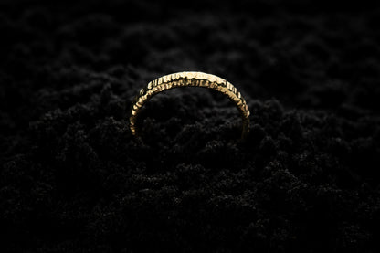 A slim, yellow gold wedding ring sits in black sand to contrast its warm, luscious glow. There are hammered striations around the band like the suns rays. It is half submerged in the sand and breaks through the surface like a golden rainbow.