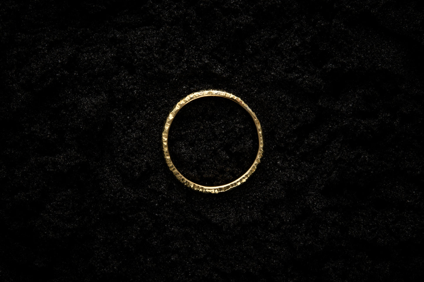 A slim, yellow gold wedding ring sits atop black sand to contrast its warm, luscious glow. There are hammered striations around the band like the suns rays. It is photographed from the top and looks like a solar eclipse.