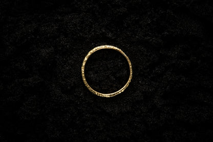 A slim, yellow gold wedding ring sits atop black sand to contrast its warm, luscious glow. There are hammered striations around the band like the suns rays. It is photographed from the top and looks like a solar eclipse.
