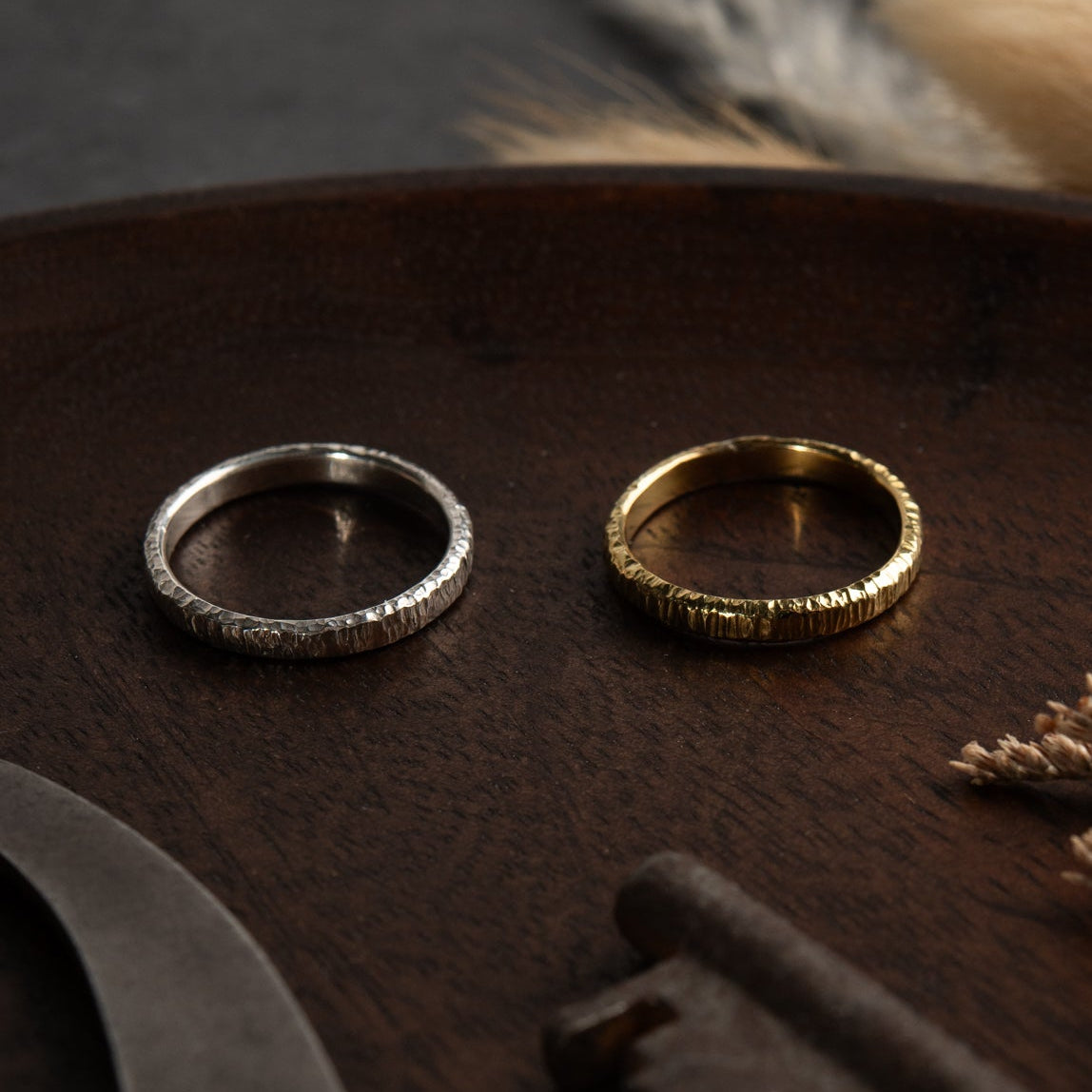 A yellow and white gold matching wedding band pair sit a wooden surface with dried plants in the background. The rings have hammered striations that glisten in the light. The inside of the bands are smooth and look comfortable.
