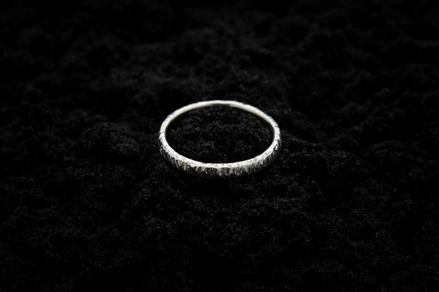 A slim, silver wedding ring sits atop black sand to contrast its bright, shiny glow. There are hammered striations around the band like the suns rays.