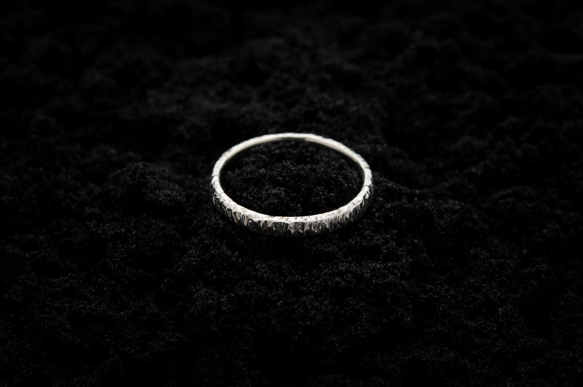 A slim, silver wedding ring sits atop black sand to contrast its bright, shiny glow. There are hammered striations around the band like the suns rays.