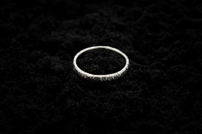 A slim, silver wedding ring sits atop black sand to contrast its bright, shiny glow. There are hammered striations around the band like the suns rays.