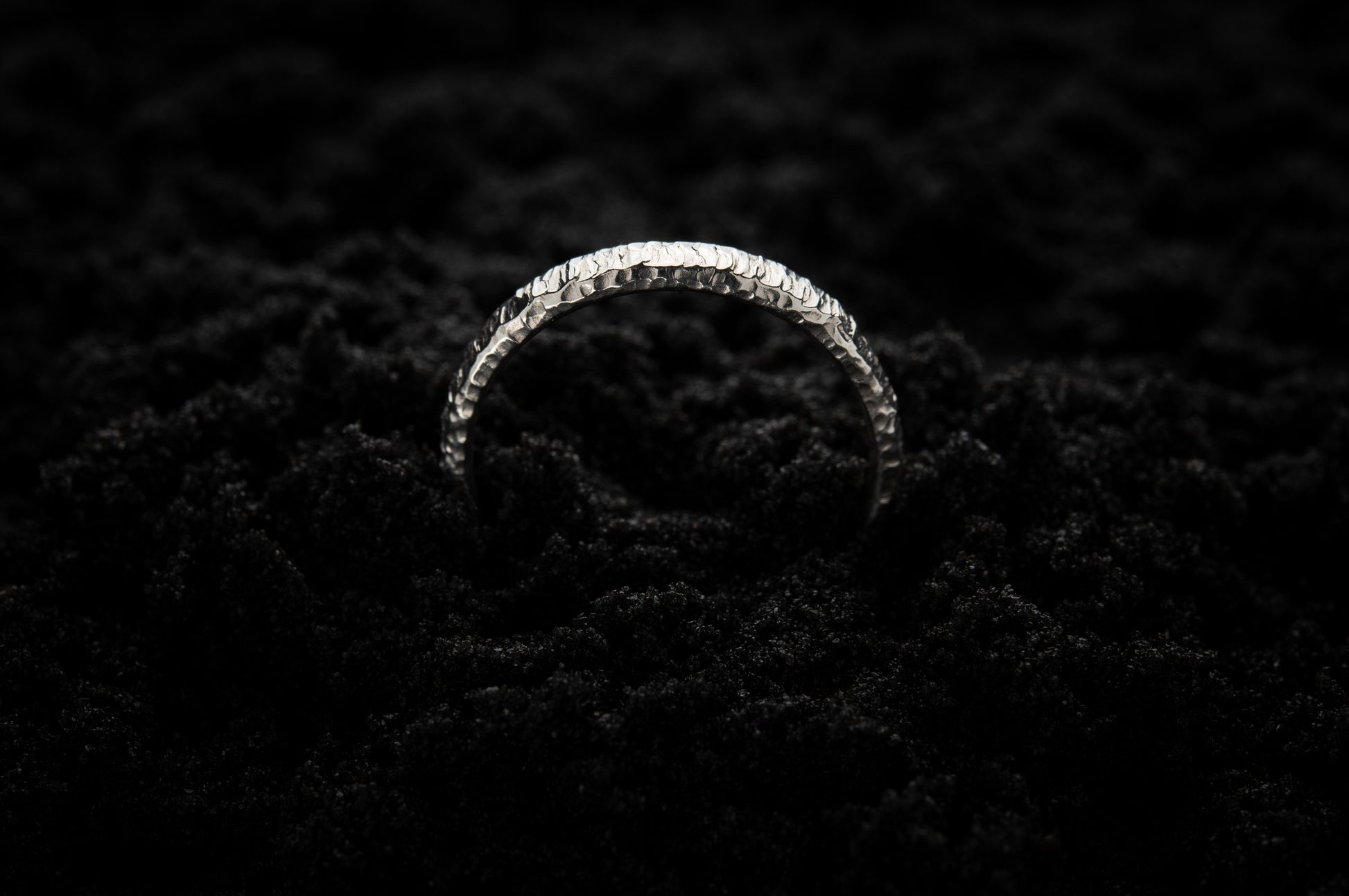 A slim, white gold wedding ring sits in black sand to contrast its bright, luscious glow. There are hammered striations around the band like the suns rays. It is half submerged in the sand and breaks through the surface like a silver rainbow.