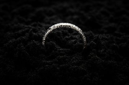 A slim, white gold wedding ring sits in black sand to contrast its bright, luscious glow. There are hammered striations around the band like the suns rays. It is half submerged in the sand and breaks through the surface like a silver rainbow.