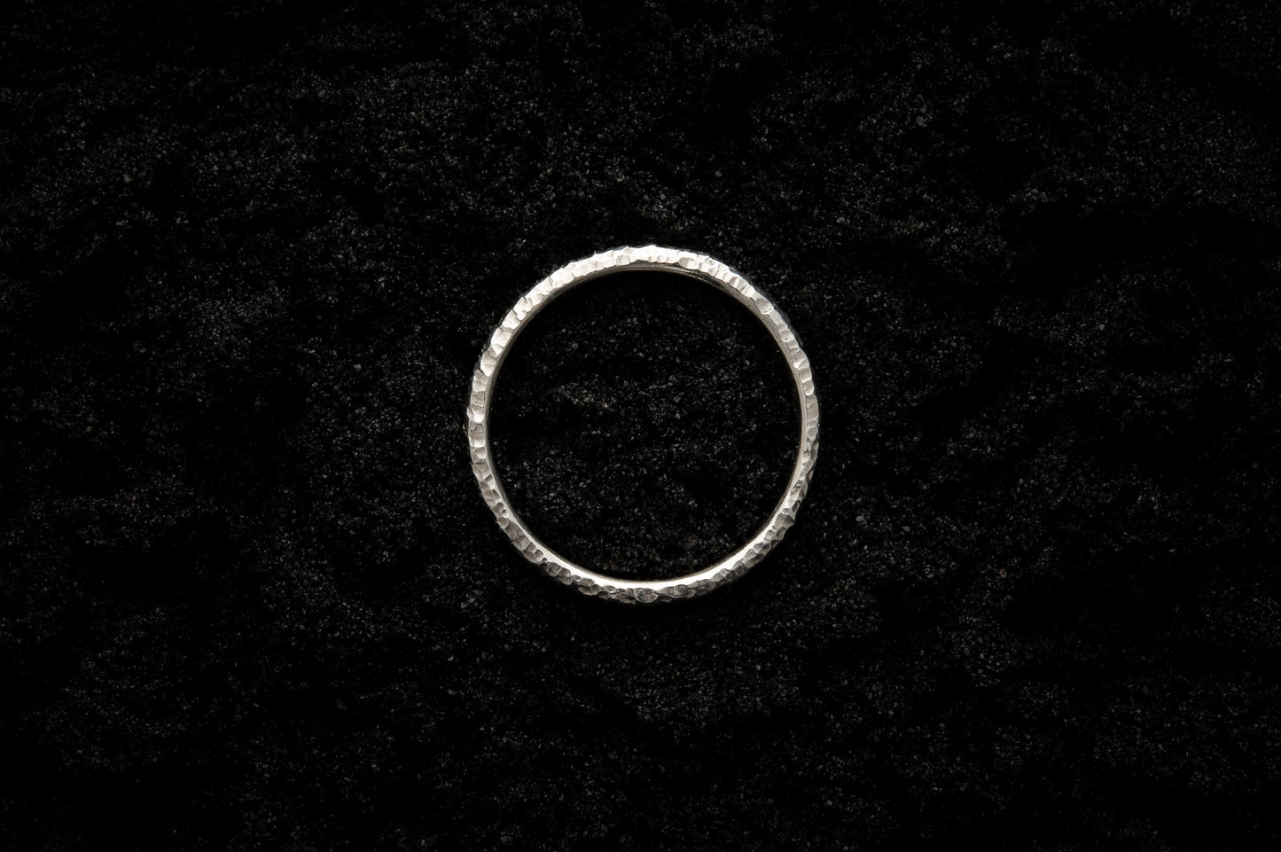 A slim, white gold wedding ring sits atop black sand to contrast its bright, shining glow. There are hammered striations and its photographed from above, reminiscent of a lunar eclipse.