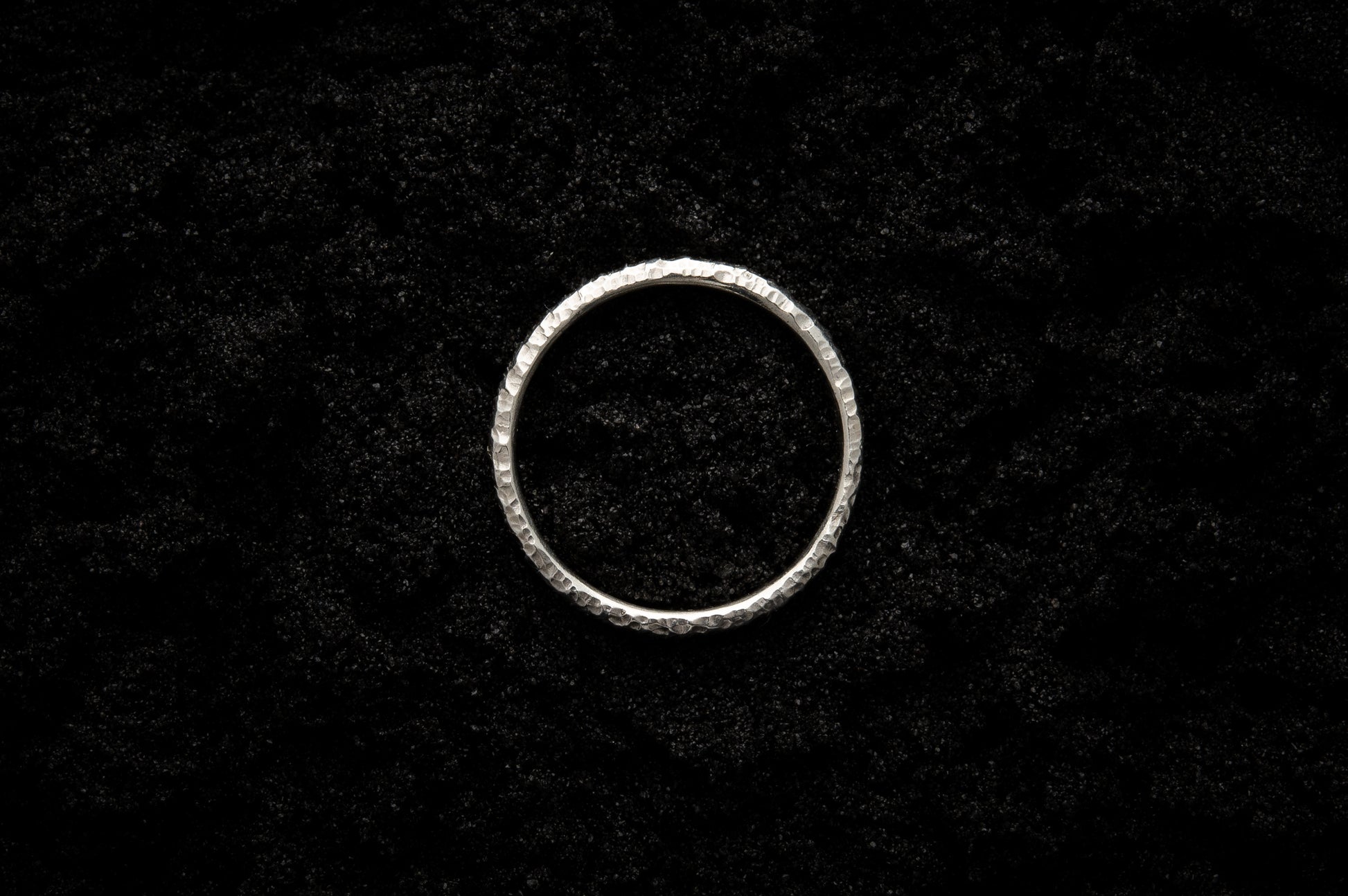 A slim, white gold wedding ring sits atop black sand to contrast its bright, shining glow. There are hammered striations and its photographed from above, reminiscent of a lunar eclipse.