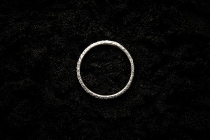 A slim, white gold wedding ring sits atop black sand to contrast its bright, shining glow. There are hammered striations and its photographed from above, reminiscent of a lunar eclipse.