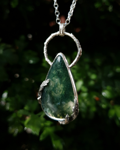 A silver pendant hangs from a chain in front of green, out-of-focus background. It is comprised of a large, smooth, teardrop-shaped moss agate bezel-set in silver with a halo-like bail made from the solid-silver casting of a real twig. There are solid silver castings of cypress-sprigs surrounding the stone too. The silver is oxidised and polished back to reveal all the rich, natural textures of the twigs. The pendant is illuminated in natural sunlight showing the beautiful inclusions of the moss agate.