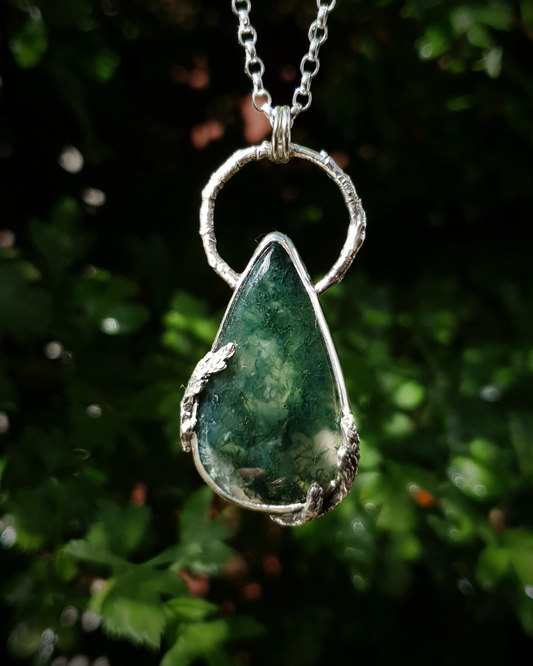 A silver pendant hangs from a chain in front of green, out-of-focus background. It is comprised of a large, smooth, teardrop-shaped moss agate bezel-set in silver with a halo-like bail made from the solid-silver casting of a real twig. There are solid silver castings of cypress-sprigs surrounding the stone too. The silver is oxidised and polished back to reveal all the rich, natural textures of the twigs. The pendant is illuminated in natural sunlight showing the beautiful inclusions of the moss agate.