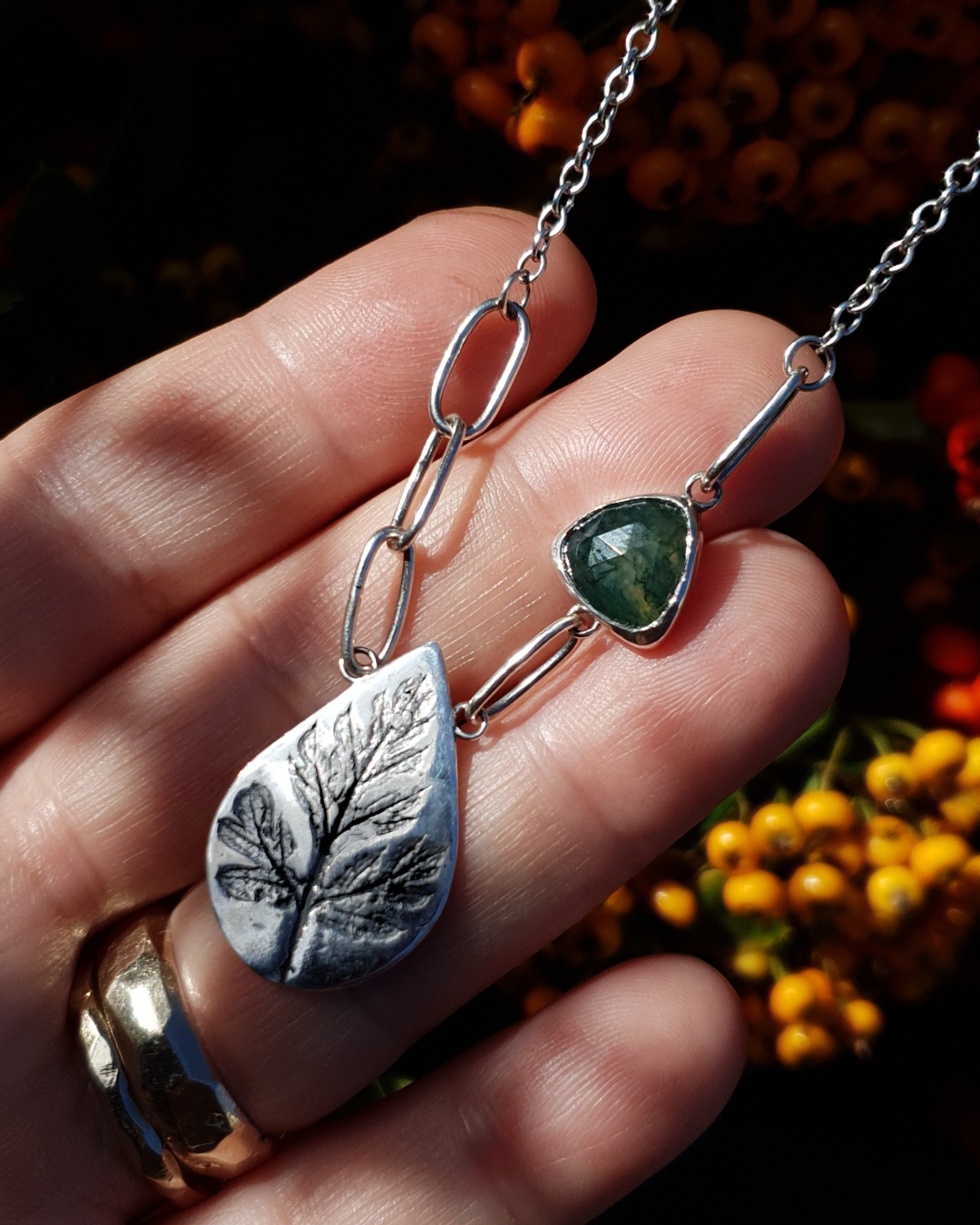 A silver necklace sits on an open palm. It's comprised of a large silver teardrop with the oxidised impression of a leaf in it reminiscent of a fossil. To the left of the pendant there are 3 large oval links which join to the rest of the chain which has much smaller links. To the right of the pendant there is 1 large oval link, then a triangular moss agate, set in silver, and then a 3rd large oval link which joins to the rest of the chain. The moss agate in the sunlight shows the beautiful green inclusions.