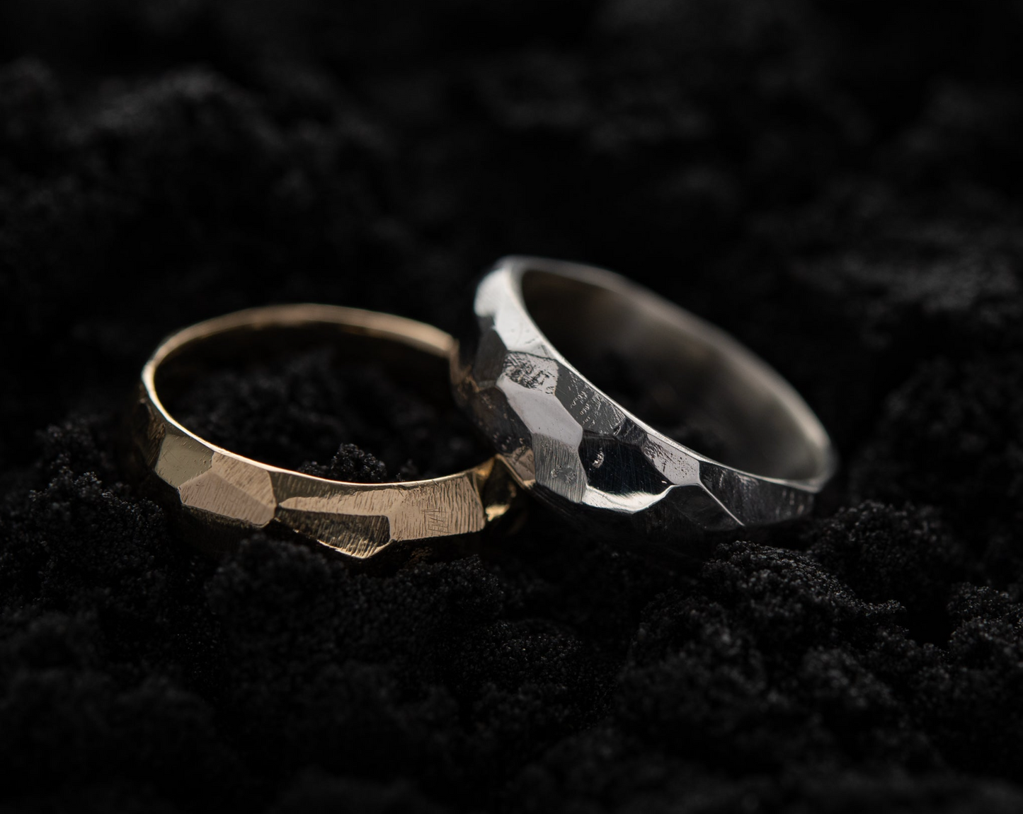 A gold and silver rough cut ring sit in black sand, the silver ring leaning against the gold one.