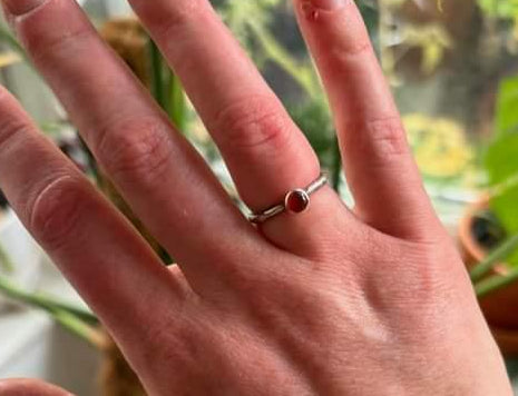 A hand wearing a solid silver ring with a small, circular, carnelian gemstone. The band of the ring is textured like a twig.
