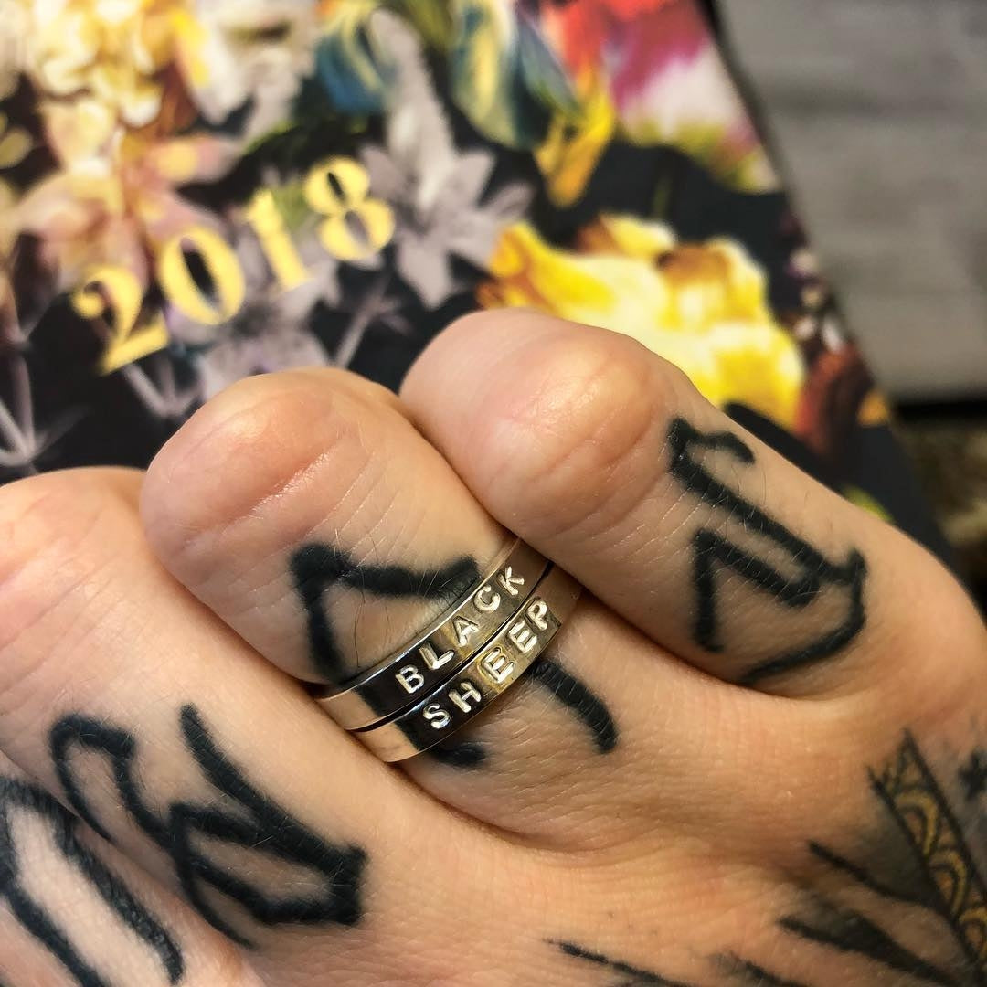 A tattooed hand wears 2 silver rings with the words "black sheep" stamped into them