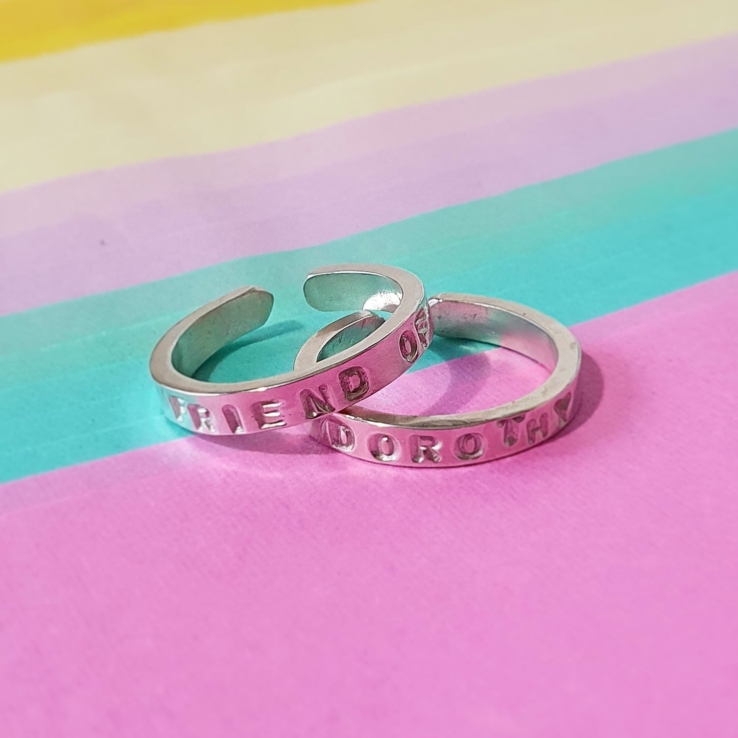 2 Silver rings sit on a colourful background. The words on the rings spell "Friend of Dorothy". The rings are left open so they are adjustable for different sizes.