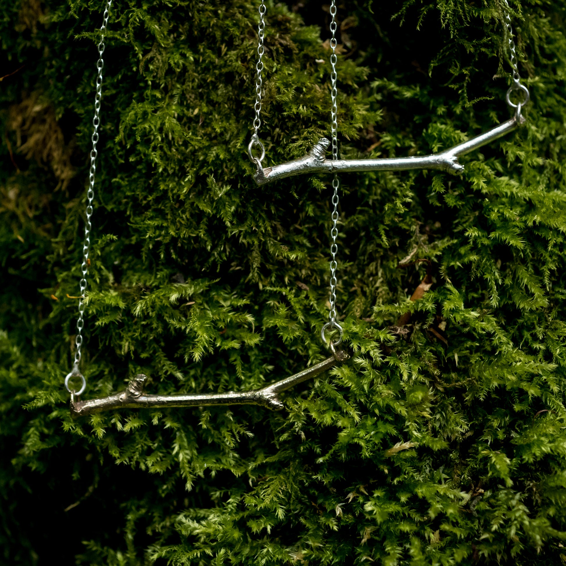 A Bronze and Silver Woodland necklace hang together against a green, mossy background