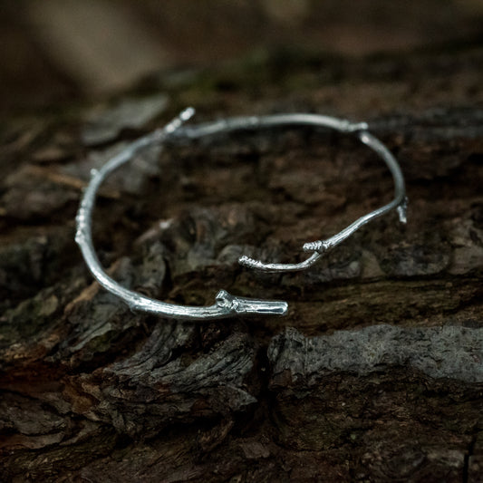 A silver twig bracelet sits on the dark bark of a fallen tree.