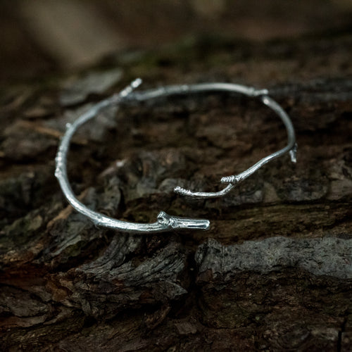 A silver twig bracelet sits on the dark bark of a fallen tree.