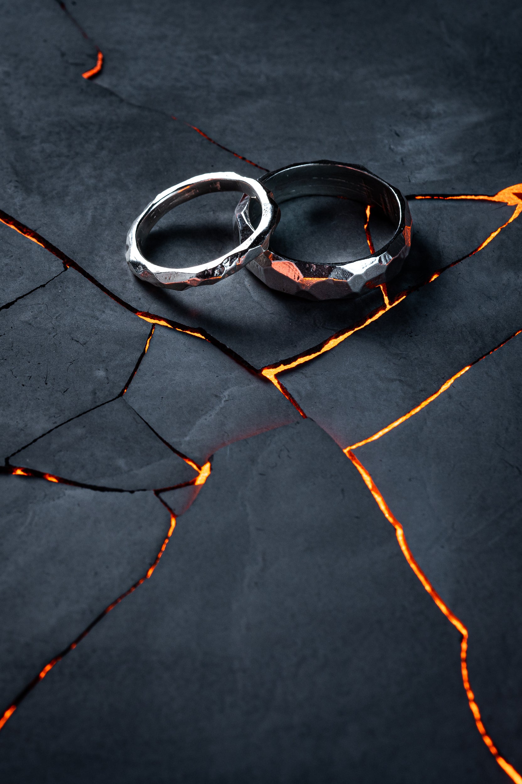 A Thin and a Thick silver ring, with rough faceted edges sit against a cracked slate background. Warm orange light glows through the cracks like lava.