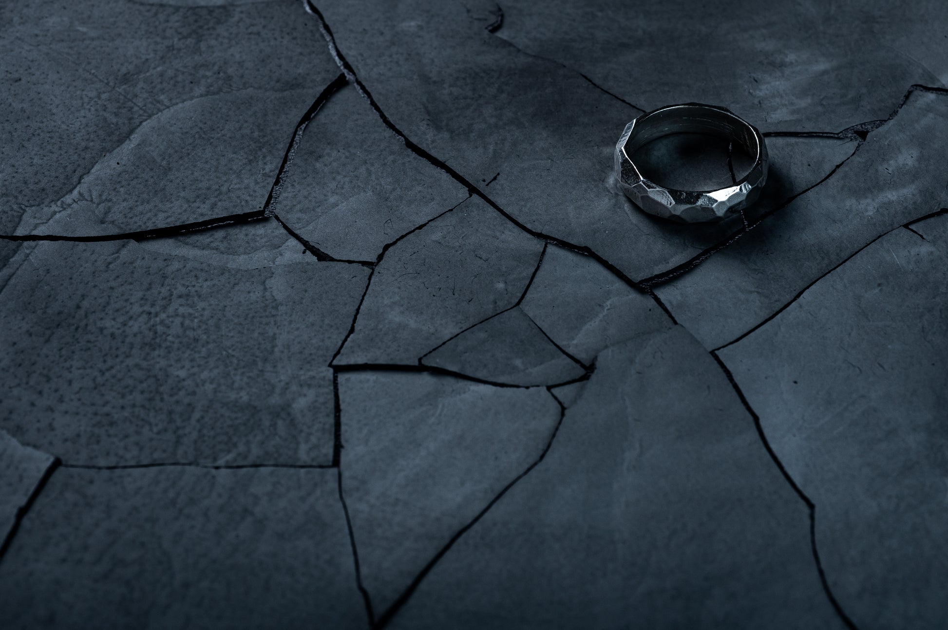 A silver, rough faceted ring sits on a cracked slate background. The lighting is dark and moody.