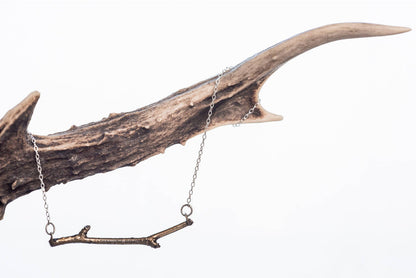A Bronce Woodland Necklace hangs from an antler on a silver chain. The background is white.
