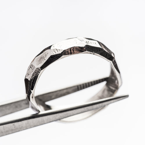 A silver, rough cut and faceted ring is held by tweezers against a crisp, white background
