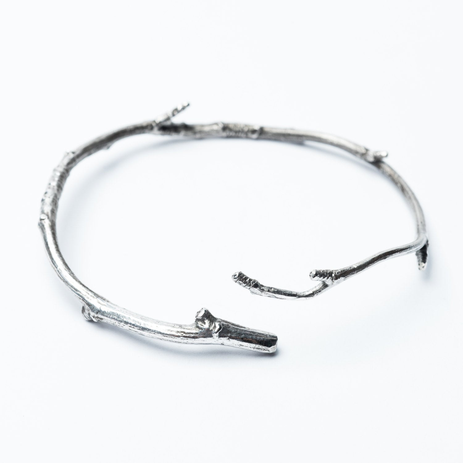 A silver bangle bracelet with a rough, natural texture, displayed on a white background. The bracelet is made from a piece of twig that has been cast in solid sterling silver.