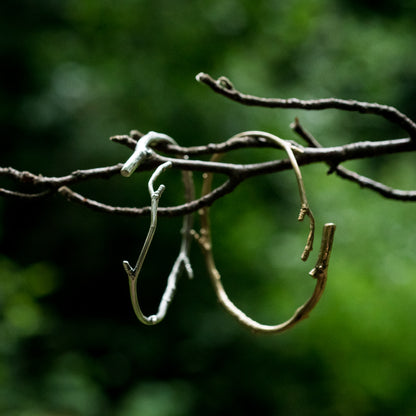 A solid silver and gold toned bracelet, cast from twigs, hang on a real twig in the foreground. the background is lush green and out of focus.